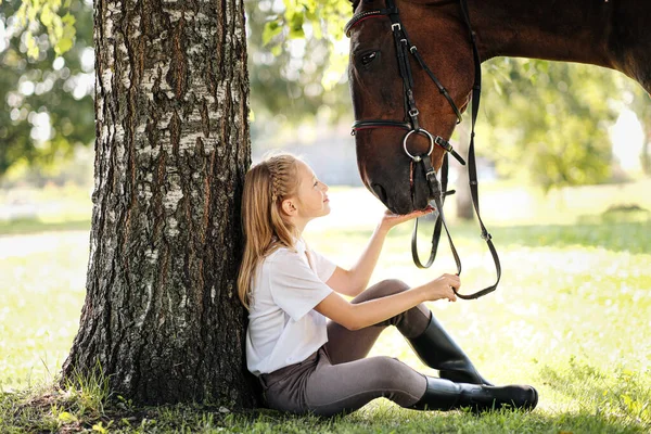 Chica jinete adolescente se sienta en un claro verde bajo un árbol. Alimenta a un caballo con una manzana y lo acaricia . — Foto de Stock