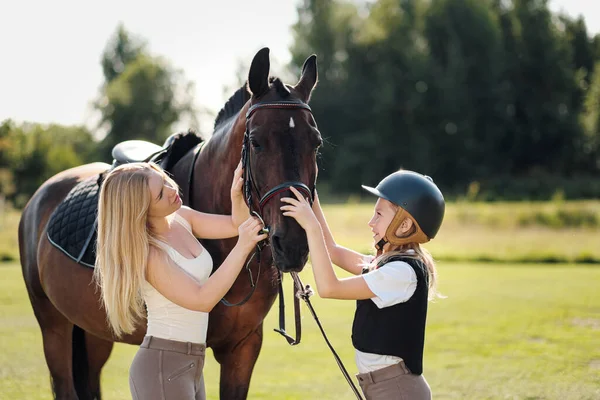 Mãe e filha pilotos e jóqueis em um campo verde abraçar um cavalo marrom . — Fotografia de Stock