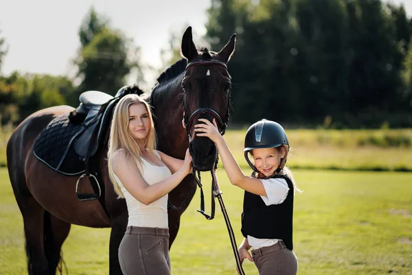 Madre e hija jinetes y jinetes en un campo verde abrazan a un caballo marrón . — Foto de Stock