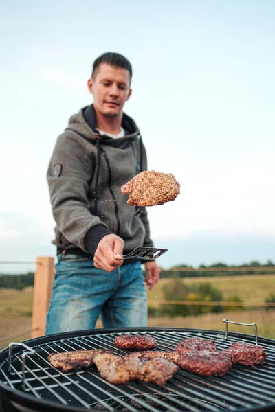 Churrasqueira. Um homem frita bifes de carne e almôndegas para hambúrgueres, vira a carne com pinças. . — Fotografia de Stock