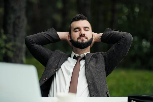 Jonge aantrekkelijke mannelijke zakenman in pak zittend aan een bureau in het bos van het natuurpark. Rekken en rusten van het werk. — Stockfoto