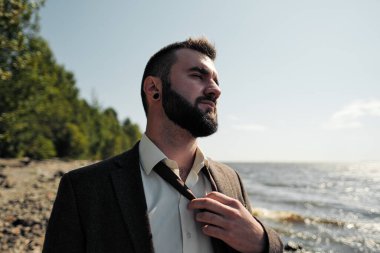Kahverengi takım elbiseli genç ve çekici bir erkek işadamı deniz kıyısında duruyor. Bağlarını çözer, bağlarını çözer ve ellerini denize kaldırır..