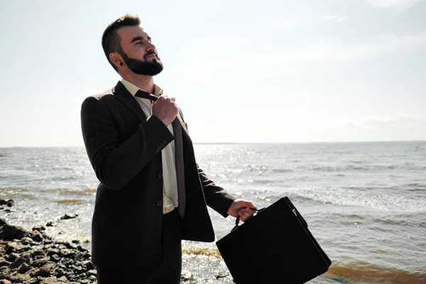 Young attractive male businessman in a brown suit with a briefcase stands on the seashore. Unfastens shirt, loosens tie and raises hands to sea.