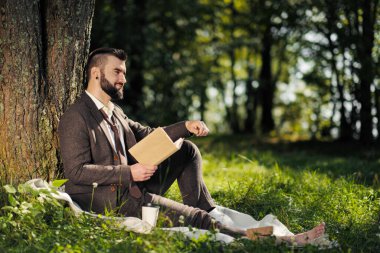 Genç ve çekici sakallı iş adamı ağacın altındaki yeşil çimlerde oturuyor ve parkta dinleniyor. Kitap oku, kahve iç. Rahatlamış, yorgun, öğle yemeği molası.