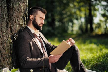 Genç ve çekici sakallı iş adamı ağacın altındaki yeşil çimlerde oturuyor ve parkta dinleniyor. Kitap oku, kahve iç. Rahatlamış, yorgun, öğle yemeği molası.