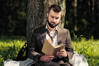 Genç ve çekici sakallı iş adamı ağacın altındaki yeşil çimlerde oturuyor ve parkta dinleniyor. Kitap oku, kahve iç. Rahatlamış, yorgun, öğle yemeği molası.