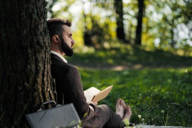 Genç ve çekici sakallı iş adamı ağacın altındaki yeşil çimlerde oturuyor ve parkta dinleniyor. Kitap oku, kahve iç. Rahatlamış, yorgun, öğle yemeği molası.