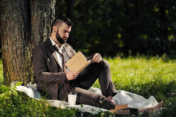 Jeune homme d'affaires barbu attrayant assis sur l'herbe verte sous l'arbre et se reposant dans le parc. Lire le livre, boire du café. Détendu, fatigué du travail, pause déjeuner . — Photo