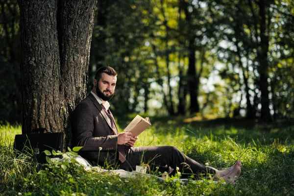 Jeune homme d'affaires barbu attrayant assis sur l'herbe verte sous l'arbre et se reposant dans le parc. Lire le livre, boire du café. Détendu, fatigué du travail, pause déjeuner . — Photo