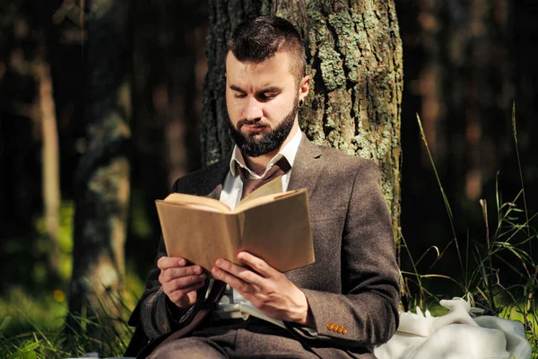 Joven hombre de negocios barbudo atractivo sentado en la hierba verde bajo el árbol y descansando en el parque. Lee el libro, bebe café. Relajado, cansado del trabajo, hora del almuerzo . —  Fotos de Stock