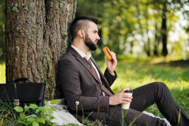 Sakallı, ağaçların altındaki yeşil çimlerde oturan ve parkta dinlenen çekici genç bir iş adamı. Kahve içer ve sandviç yer. Rahatlama, çalışmaktan yorulma, öğle yemeği molası..
