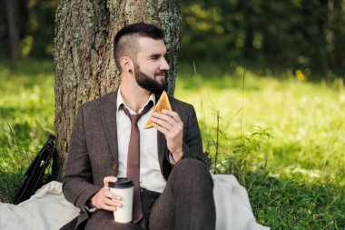 Sakallı, ağaçların altındaki yeşil çimlerde oturan ve parkta dinlenen çekici genç bir iş adamı. Kahve içer ve sandviç yer. Rahatlama, çalışmaktan yorulma, öğle yemeği molası..