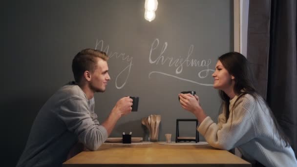 Jeune couple à la maison dans la cuisine du soir. Ils boivent du thé, rient et parlent. Une lampe jaune est allumée au-dessus de la table. Veille de Noël . — Video