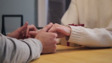 Bir çift evde, masada oturuyor. Bir erkek kadınların ellerini tutar ve onları okşar. Hediye kutusu.