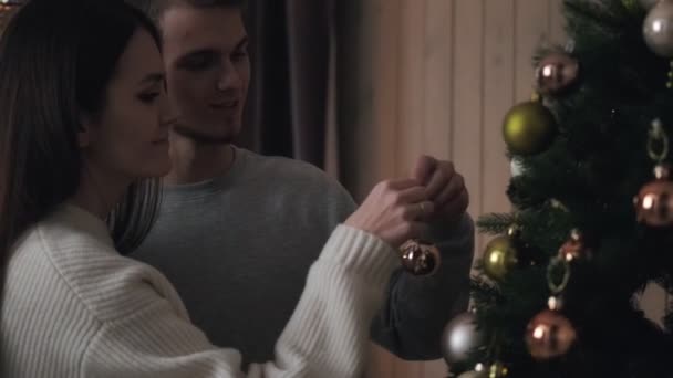 Een jong stel versiert 's avonds een kerstboom thuis. Ballen hangen, knuffelen en lachen. Kerstavond. — Stockvideo