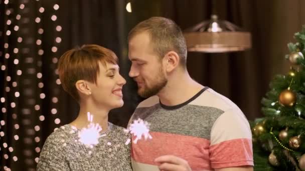 Jeune couple marié à la maison dans des vêtements décontractés, un homme et une femme tirant scintille à côté d'un arbre de Noël. Célébrez les vacances . — Video