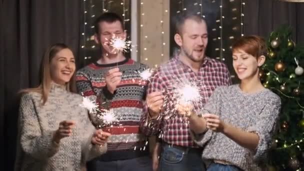 Un grupo de amigos en suéteres, cuatro personas, en el fondo de un árbol de Navidad, queman bengalas . — Vídeo de stock