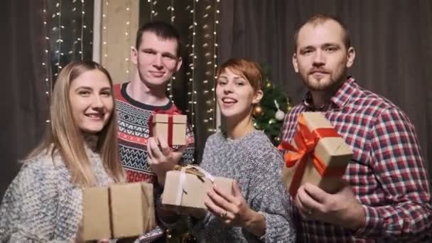 Un groupe d'amis en chandails, quatre personnes, devant un sapin de Noël, tiennent des boîtes cadeaux dans leurs mains riant et s'amusant . — Video