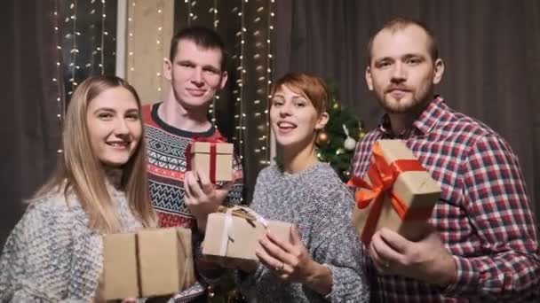 Un grupo de amigos en suéteres, cuatro personas, frente a un árbol de Navidad, llevan cajas de regalo en sus manos riendo y divirtiéndose . — Vídeo de stock