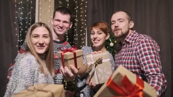 Skupina přátel ve svetrech, čtyři lidé, před vánočním stromečkem, drží dárkové krabice v rukou se smíchem a baví se. — Stock video