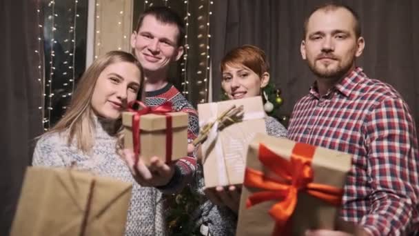 Un grupo de amigos en suéteres, cuatro personas, frente a un árbol de Navidad, llevan cajas de regalo en sus manos riendo y divirtiéndose . — Vídeo de stock