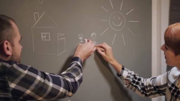 Young married couple in casual clothes, man and woman, sitting at home in the kitchen at the table. Draw their dreams with chalk on the wall. — Stock Video