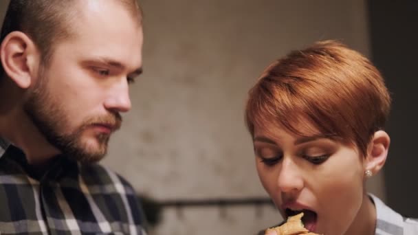 Jeune couple marié en vêtements décontractés, homme et femme mangeant des sandwichs à la maison dans la cuisine. Lumière du soir des lampes jaunes . — Video