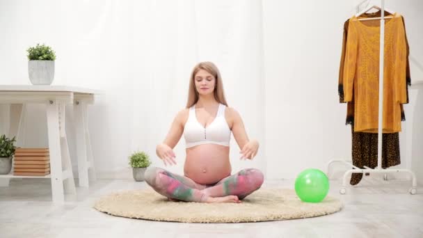 En ung vacker gravid vit kvinna sitter på husets mjuka matta. Han är engagerad i fitness och gör övningar. Sportkläder. — Stockvideo