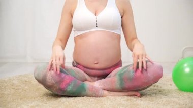 Genç, güzel, hamile, beyaz bir kadın evin yumuşak halısında oturuyor. Spor yapıyor, spor yapıyor, spor yapıyor. Spor kıyafetleri..