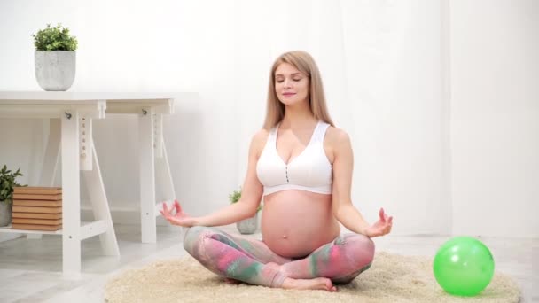 若い美しい妊婦の白人女性が家の柔らかいカーペットの上に座っています。彼はフィットネスと運動に従事しています.スポーツウェア. — ストック動画