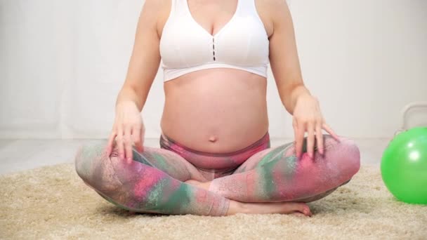 Молодая красивая беременная белая женщина сидит на мягком ковре дома. Занимается фитнесом и упражнениями. Спортивная одежда . — стоковое видео