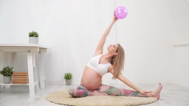 Uma jovem mulher branca grávida bonita senta-se no tapete macio da casa. Envolve-se na aptidão e fazendo exercícios. Roupa de desporto . — Vídeo de Stock