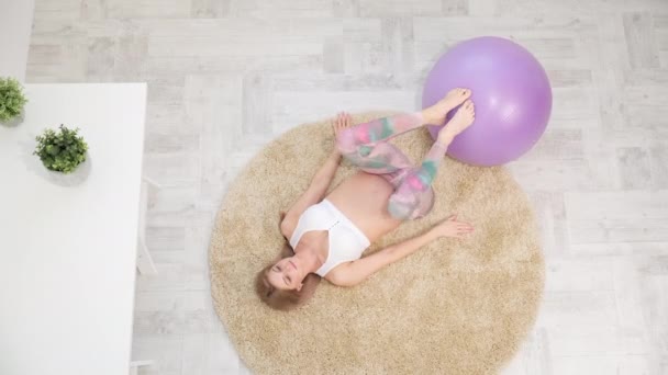 A young beautiful pregnant Caucasian woman sits on the soft carpet of the house. He is engaged in fitness and doing exercises. Sportswear. — Stock Video