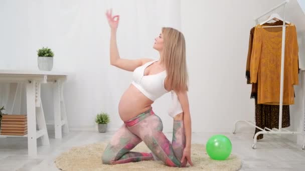 Eine junge schöne schwangere Kaukasierin sitzt auf dem weichen Teppich des Hauses. Er betreibt Fitness und macht Übungen. Sportbekleidung. — Stockvideo