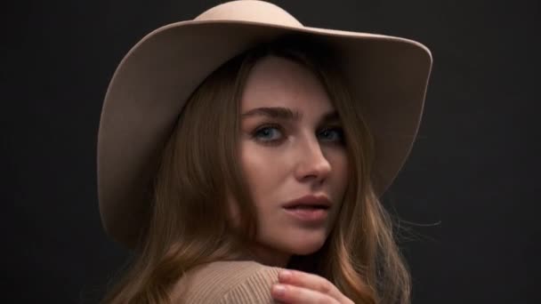Mladá krásná elegantní žena v béžovém svetru a klobouku s poli pózuje na černém pozadí. Detailní záběr na tvář. — Stock video