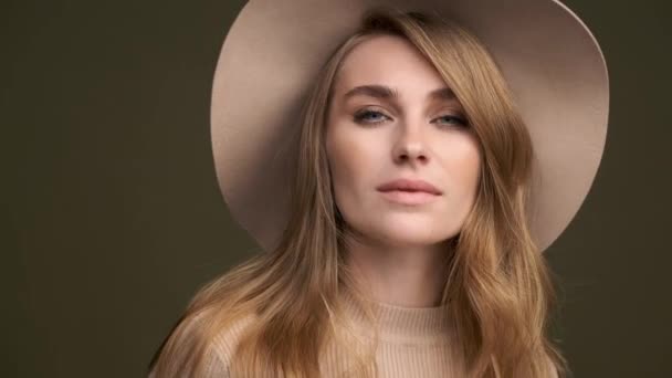 Een jonge mooie elegante vrouw in een beige trui en fedora met velden vormt tegen een groene olijf achtergrond. Een close-up van het gezicht. — Stockvideo