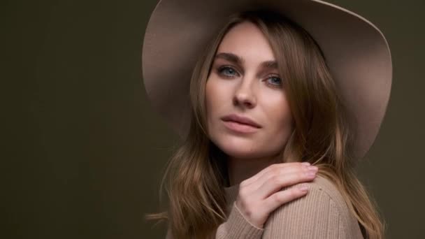 Uma jovem mulher bonita e elegante em um suéter bege e fedora com campos posa contra um fundo verde azeitona. Um close-up do rosto . — Vídeo de Stock