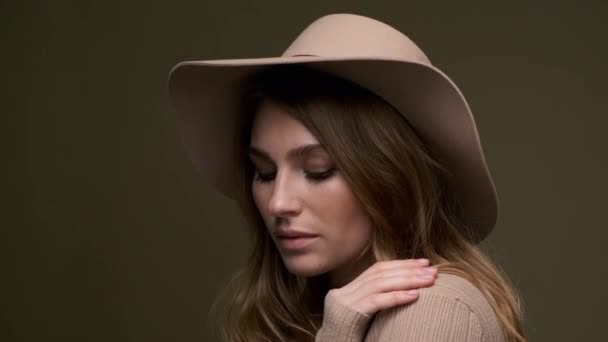 Uma jovem mulher bonita e elegante em um suéter bege e fedora com campos posa contra um fundo verde azeitona. Um close-up do rosto . — Vídeo de Stock