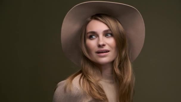 A young beautiful elegant woman in a beige sweater and fedora with fields poses against a green olive background. A close-up of the face. — Stock Video