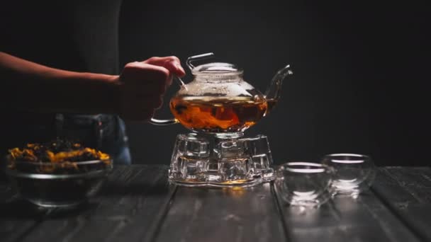 Tea ceremony. The womans hand pours hot black tea with flowers from a transparent teapot into glass piala cups. — Stock Video