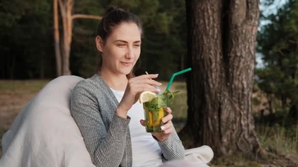 Eine junge attraktive Frau entspannt sich in der Natur. Trinkt einen Cocktail und kommuniziert mit Freunden. — Stockvideo