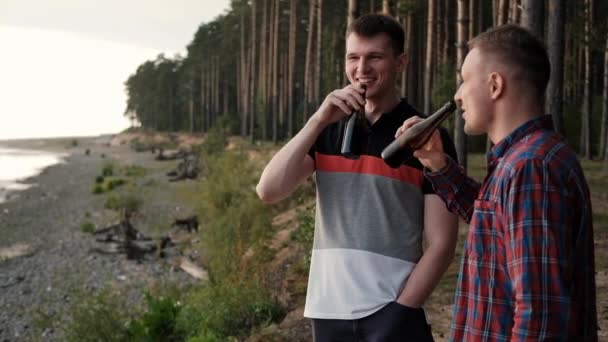 Deux jeunes hommes boivent de la bière dans des bouteilles et se détendent à l'extérieur. Un week-end dans la nature, une rencontre d'amis . — Video