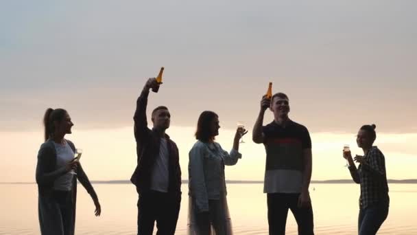Impreza przyjaciół na plaży o zachodzie słońca. Kobiety i mężczyźni dobrze się bawią, piją piwo i wino, tańczą i śmieją. — Wideo stockowe