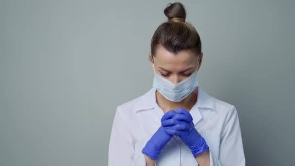 Μια γυναίκα γιατρός με λευκή ρόμπα, μάσκα και γάντια. Κοίτα από κοντά. Ο γιατρός κλαίει και προσεύχεται. Δάκρυα στα μάτια. Πανδημία και επιδημία ιών. Ιός του Coronavirus covid-19. — Αρχείο Βίντεο