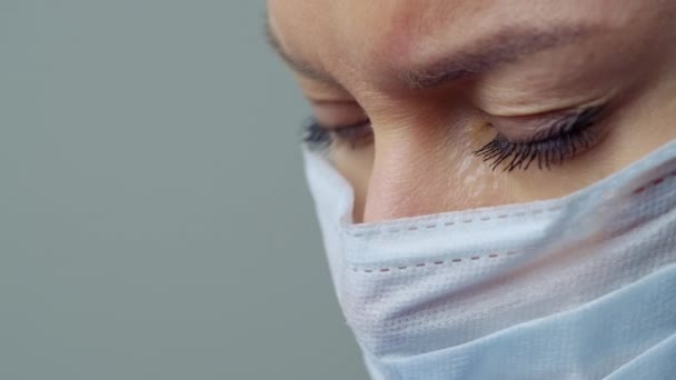 Een vrouwelijke arts therapeut in een wit gewaad, masker en handschoenen. Gezicht van dichtbij. De dokter huilt en bidt. Tranen in de ogen. Pandemie en virusepidemie. Coronavirus covid-19. — Stockvideo