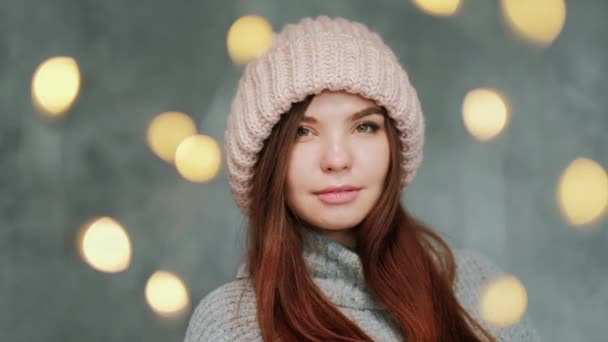 Gri kazaklı ve gri arka planda pembe örülü şapka takan genç bir kadının portresi. Bulanık ışıklar. — Stok video