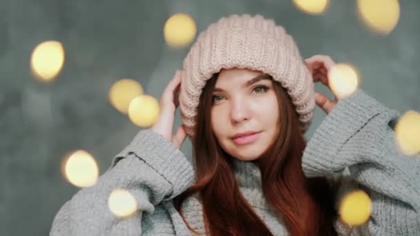 Retrato de una mujer joven en un suéter gris y un sombrero de punto rosa sobre un fondo gris. Luces borrosas . — Vídeos de Stock