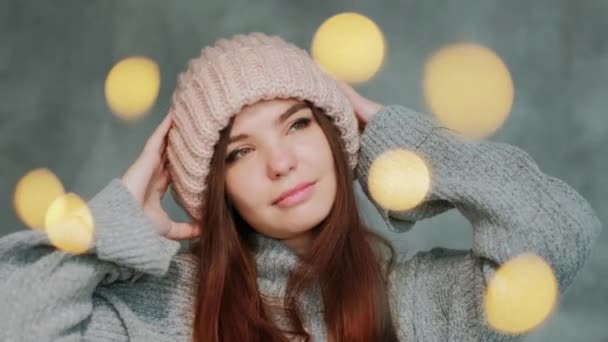 Portret van een jonge vrouw in een grijze trui en roze gebreide hoed op een grijze achtergrond. Wazige lichten. — Stockvideo