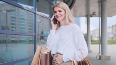 Beyaz kazaklı ve spor ayakkabılı güzel bir sarışın kadın dükkandan gelen paketlerle caddede yürüyor. Hamile kadın alışverişi..