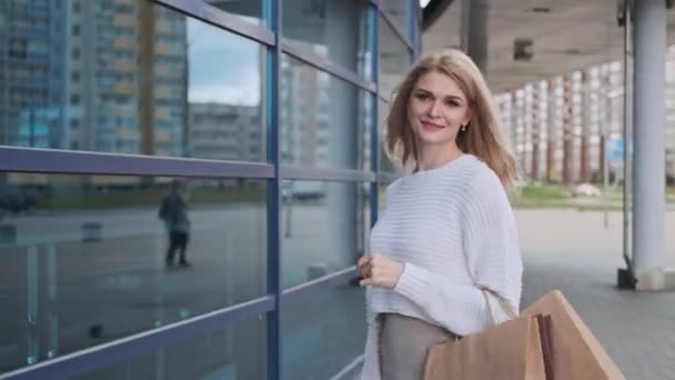 Una joven rubia hermosa en un suéter blanco y zapatillas de deporte camina por la calle con paquetes de la tienda . — Vídeo de stock